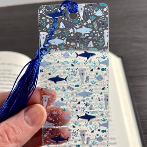 May include: A rectangular, clear acrylic bookmark featuring a repeating pattern of various sharks, including hammerheads, in shades of blue, white, and teal, interspersed with small sea life and coral motifs, finished with a blue tassel.