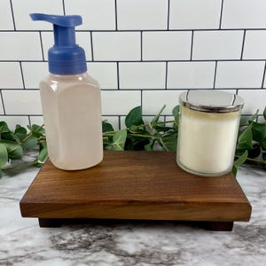 May include: A wooden tray holds a light pink soap dispenser with a blue pump and a glass jar candle with a silver lid. The tray is dark brown and rectangular.