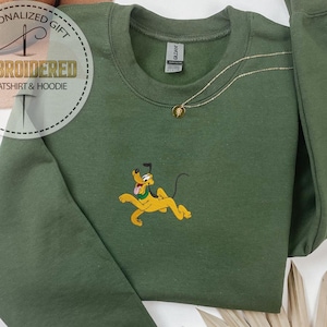 Peut inclure: Un sweat-shirt vert olive &agrave; col rond et manches longues. Le sweat-shirt pr&eacute;sente une image brod&eacute;e de Pluto, un chien de dessin anim&eacute; jaune. Un collier en or avec un pendentif est visible. Les mots "EMBROIDERED SWEATSHIRT & HOODIE" sont imprim&eacute;s sur une &eacute;tiquette circulaire.