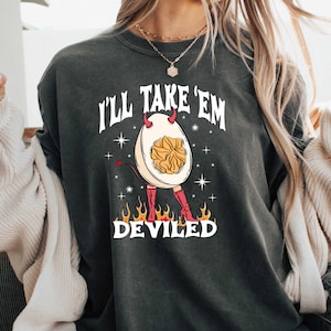 May include: Dark grey t-shirt with the text "I'll Take 'Em Deviled". The graphic features a deviled egg character with horns, a tail, and boots, surrounded by flames. The shirt is a casual wear item.