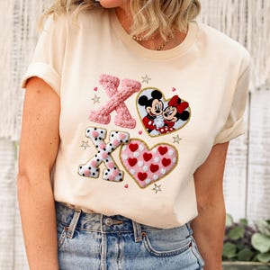 May include: Cream-colored t-shirt with embroidered Valentine's Day design. Features pink and black X's, a heart with red hearts, and a Mickey and Minnie Mouse heart. The shirt is paired with blue jeans.