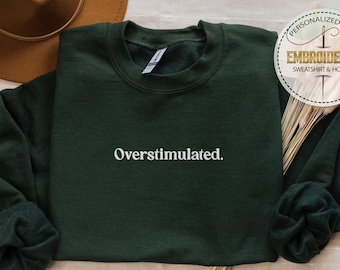 May include: A dark green sweatshirt with the text "Overstimulated." embroidered in white on the front. The sweatshirt is folded and laid flat on a light beige surface.