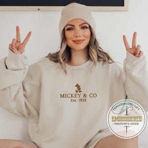 Puede incluir: Una sudadera beige con una silueta de Mickey Mouse marr&oacute;n y el texto "Mickey & Co. Est. 1928".