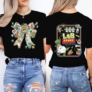May include: Black t-shirt featuring a colorful bow design with various cartoon characters. The back of the shirt has a rectangular graphic with the text "Lab Story" and a dinosaur in a lab coat. The shirt is paired with blue jeans.