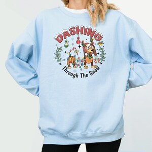 May include: Pale blue sweatshirt with a Christmas-themed graphic. The graphic includes a cartoon dog and toy soldier, with the words "Dashing Through The Snow". The sweatshirt is a plain light blue colour.