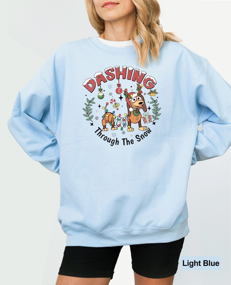 May include: Pale blue sweatshirt with a Christmas-themed graphic. The graphic includes a cartoon dog and toy soldier, with the words "Dashing Through The Snow". The sweatshirt is a plain light blue colour.