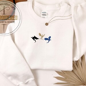 May include: White embroidered sweatshirt featuring three dragon designs in black, gold, and blue. The text "PERSONALIZED GIFT" and "EMBROIDERED SWEATSHIRT & HOODIE" are on a circular graphic. A gold necklace with a pendant is also visible.