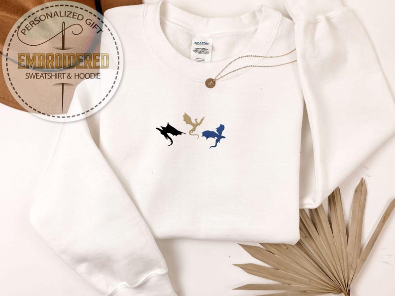 May include: White embroidered sweatshirt featuring three dragon designs in black, gold, and blue. The text "PERSONALIZED GIFT" and "EMBROIDERED SWEATSHIRT & HOODIE" are on a circular graphic. A gold necklace with a pendant is also visible.