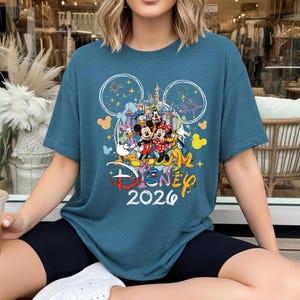 May include: Teal blue t-shirt featuring a colorful Disney graphic with Mickey and Minnie Mouse, Donald Duck, and Goofy. The design includes the text "Disney 2026" and fireworks. The shirt has a crew neck and short sleeves.
