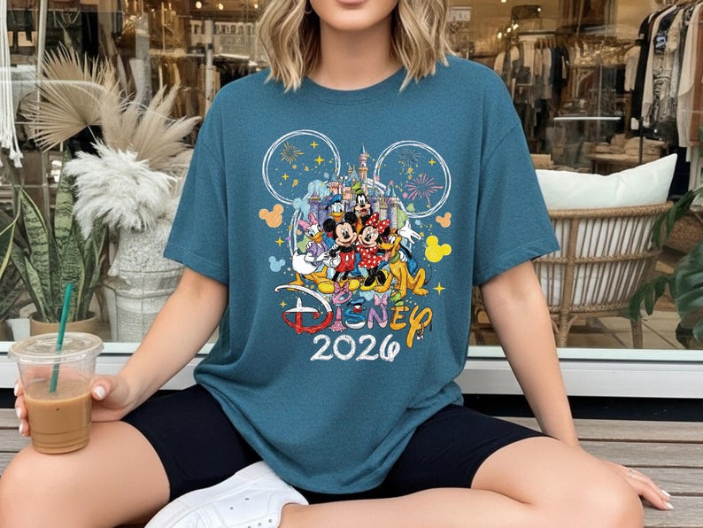 May include: Teal blue t-shirt featuring a colorful Disney graphic with Mickey and Minnie Mouse, Donald Duck, and Goofy. The design includes the text "Disney 2026" and fireworks. The shirt has a crew neck and short sleeves.