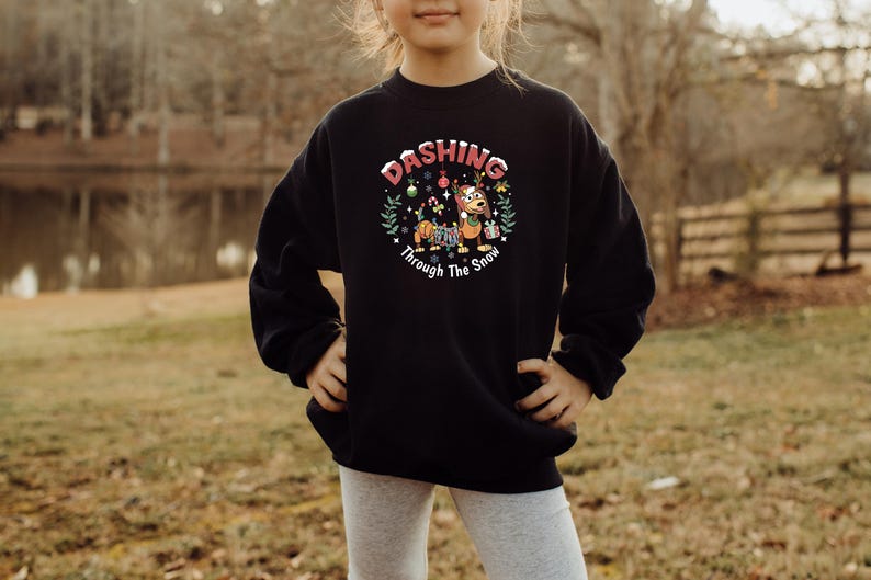 May include: Black sweatshirt with a festive graphic. The design features a dachshund dog wearing a scarf and holding a gift, surrounded by Christmas decorations and the text "Dashing Through The Snow".