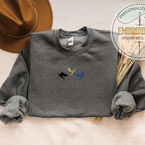 May include: A charcoal grey sweatshirt featuring embroidered dragon motifs in black, gold, and blue. The garment has a crew neck and long sleeves. The image also shows a brown hat and a tag that reads "Embroidered Sweatshirt & Hoodie".
