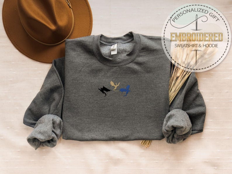 May include: A charcoal grey sweatshirt featuring embroidered dragon motifs in black, gold, and blue. The garment has a crew neck and long sleeves. The image also shows a brown hat and a tag that reads "Embroidered Sweatshirt & Hoodie".