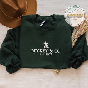 Puede incluir: Una sudadera verde oscuro con una silueta blanca de Mickey Mouse y el texto "Mickey & Co. Est. 1928".