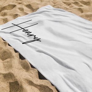 May include: White beach towel with the name "Henry" in black script. The towel is lying on a sandy surface. The name is written in a stylish, modern font.