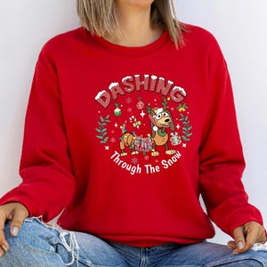 May include: A red sweatshirt with a Christmas-themed graphic. The design features a cartoon dog with antlers and fairy lights, and the text "Dashing Through The Snow". The sweatshirt is a festive item of clothing.