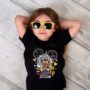May include: A black t-shirt featuring a colorful Disney graphic with Mickey Mouse, Minnie Mouse, and other characters. The shirt includes the text "Disney 2026" in a playful font. The child is wearing yellow sunglasses.
