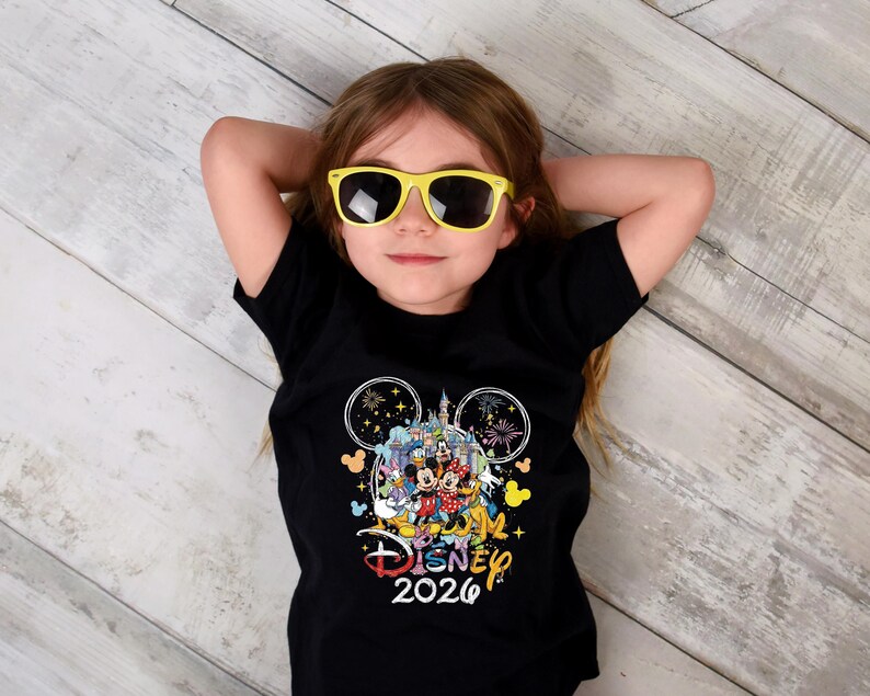 May include: A black t-shirt featuring a colorful Disney graphic with Mickey Mouse, Minnie Mouse, and other characters. The shirt includes the text "Disney 2026" in a playful font. The child is wearing yellow sunglasses.