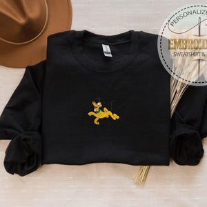 Peut inclure: Sweat-shirt noir avec un motif Pluto brod&eacute;. Le motif repr&eacute;sente Pluto en jaune, vert et noir. Le sweat-shirt a des manches longues et un col rond. L'image comprend &eacute;galement un chapeau marron et le texte "Sweat-shirt & Hoodie brod&eacute;".