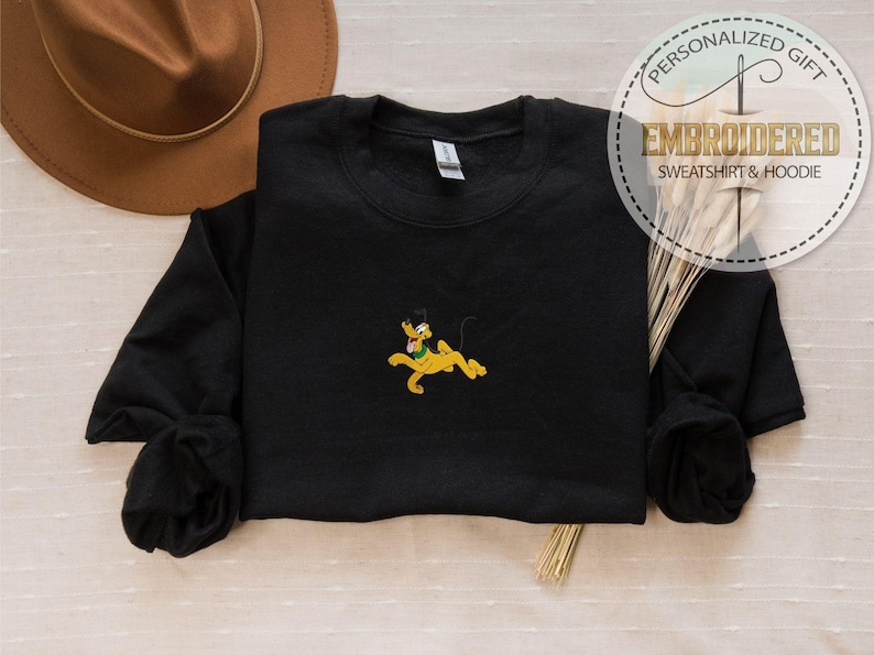 Peut inclure: Sweat-shirt noir avec un motif Pluto brod&eacute;. Le motif repr&eacute;sente Pluto en jaune, vert et noir. Le sweat-shirt a des manches longues et un col rond. L'image comprend &eacute;galement un chapeau marron et le texte "Sweat-shirt & Hoodie brod&eacute;".