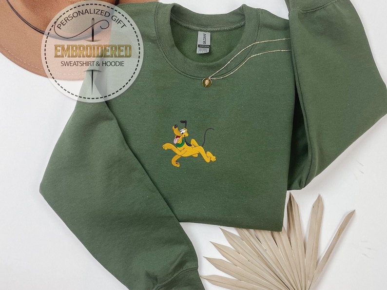 Peut inclure: Un sweat-shirt vert olive &agrave; col rond et manches longues. Le sweat-shirt pr&eacute;sente une image brod&eacute;e de Pluto, un chien de dessin anim&eacute; jaune. Un collier en or avec un pendentif est visible. Les mots "EMBROIDERED SWEATSHIRT & HOODIE" sont imprim&eacute;s sur une &eacute;tiquette circulaire.