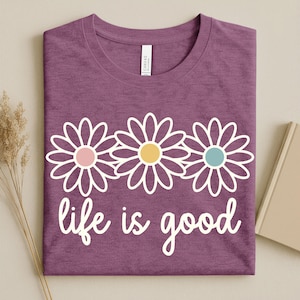 May include: A purple t-shirt featuring a white daisy design and the words "life is good" in white script. The daisies have pink, yellow, and blue centres. The shirt is neatly folded.