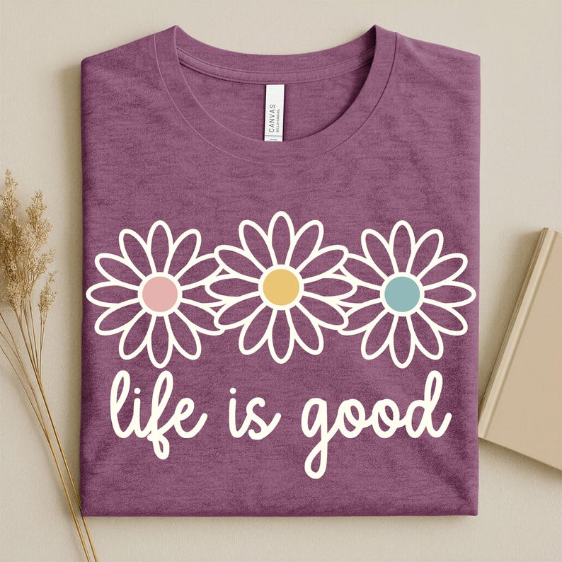 May include: A purple t-shirt featuring a white daisy design and the words "life is good" in white script. The daisies have pink, yellow, and blue centres. The shirt is neatly folded.
