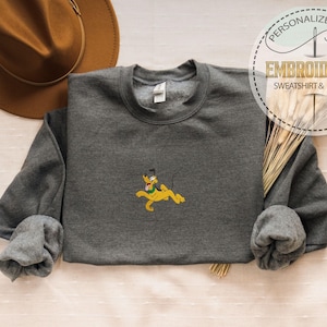 Peut inclure: Un sweat-shirt gris anthracite avec une broderie du personnage de dessin anim&eacute; Pluto en jaune. Le sweat-shirt a des manches longues et un col rond. Un chapeau marron est en arri&egrave;re-plan, avec une &eacute;tiquette indiquant "Cadeau Personnalis&eacute;, Sweat-shirt & Sweat &agrave; capuche brod&eacute;."