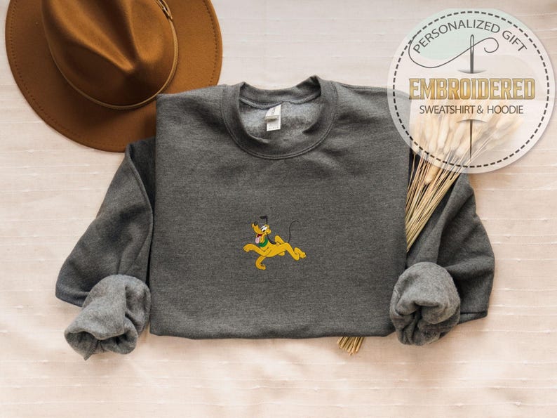 Peut inclure: Un sweat-shirt gris anthracite avec une broderie du personnage de dessin anim&eacute; Pluto en jaune. Le sweat-shirt a des manches longues et un col rond. Un chapeau marron est en arri&egrave;re-plan, avec une &eacute;tiquette indiquant "Cadeau Personnalis&eacute;, Sweat-shirt & Sweat &agrave; capuche brod&eacute;."