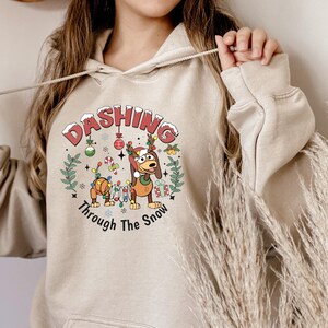 May include: Beige hoodie featuring a Christmas design. The graphic shows a cartoon dog with a Santa hat and antlers, festive decorations, and the words "Dashing Through The Snow".