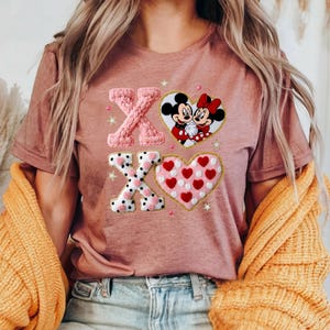 May include: Dusty rose t-shirt featuring an embroidered design with the letters "XO", a heart with Mickey and Minnie Mouse, and a heart filled with red hearts. The design includes white polka dots and star accents.