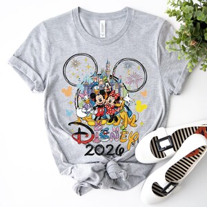 May include: A gray t-shirt featuring a colorful graphic of Disney characters, including Mickey and Minnie Mouse, in front of a castle. The text "Disney 2026" is below the graphic. White flats with black stripes are next to the shirt.