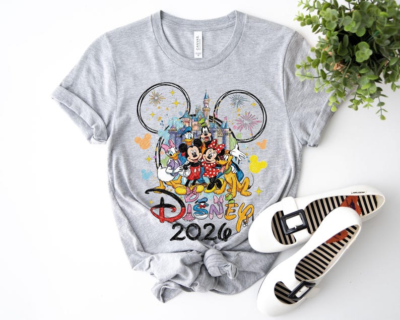May include: A gray t-shirt featuring a colorful graphic of Disney characters, including Mickey and Minnie Mouse, in front of a castle. The text "Disney 2026" is below the graphic. White flats with black stripes are next to the shirt.