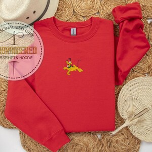 Peut inclure: Un sweat-shirt rouge avec un motif Pluto brod&eacute;. Le sweat-shirt a un col rond et des manches longues. L'image comprend &eacute;galement un chapeau de paille et un &eacute;ventail d&eacute;coratif.