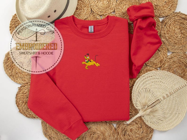 Peut inclure: Un sweat-shirt rouge avec un motif Pluto brod&eacute;. Le sweat-shirt a un col rond et des manches longues. L'image comprend &eacute;galement un chapeau de paille et un &eacute;ventail d&eacute;coratif.