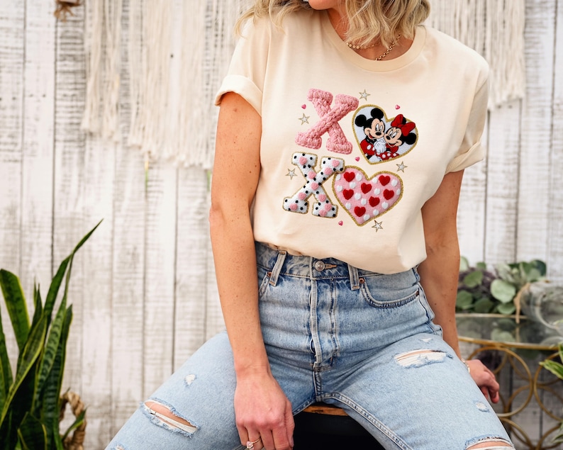 May include: Cream-colored t-shirt with embroidered Valentine's Day design. Features pink and black X's, a heart with red hearts, and a Mickey and Minnie Mouse heart. The shirt is paired with blue jeans.