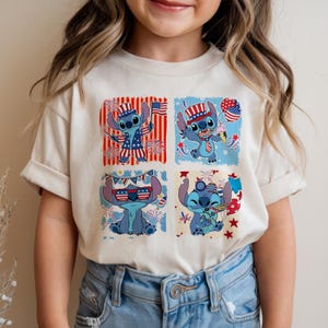 Puede incluir: Una camiseta color crema con un dise&ntilde;o de cuatro paneles de un personaje de dibujos animados azul con atuendo patri&oacute;tico. Cada panel muestra al personaje con elementos de la bandera estadounidense, fuegos artificiales y helado, celebrando el D&iacute;a de la Independencia.