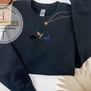 May include: Black sweatshirt featuring embroidered dragon silhouettes in black, gold, and blue. A gold necklace with a pendant is draped across the sweatshirt. The image includes a circular logo that reads "PERSONALIZED GIFT EMBROIDERED SWEATSHIRT & HOODIE".