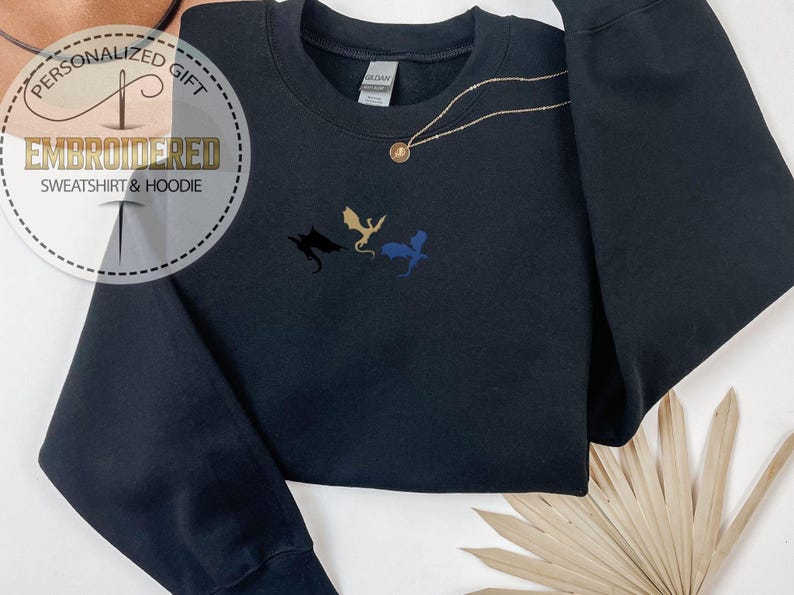May include: Black sweatshirt featuring embroidered dragon silhouettes in black, gold, and blue. A gold necklace with a pendant is draped across the sweatshirt. The image includes a circular logo that reads "PERSONALIZED GIFT EMBROIDERED SWEATSHIRT & HOODIE".