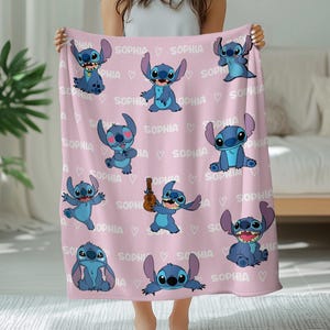 K&ouml;nnte beinhalten: Eine hellrosa Fleecedecke mit einem sich wiederholenden Muster der blauen Figur Stitch in verschiedenen Posen, durchsetzt mit dem Namen Sophia und kleinen wei&szlig;en Herzsymbolen, die in wei&szlig;er Schrift auf der Stoffoberfl&auml;che gedruckt sind.