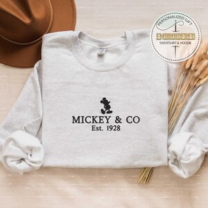 Puede incluir: Una sudadera gris con una silueta negra de Mickey Mouse y el texto "Mickey & Co. Est. 1928".