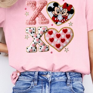 May include: Pink t-shirt featuring the letters "XO" in a knitted style, along with a heart-shaped design with Mickey and Minnie Mouse. The shirt also includes a heart-shaped design with red hearts and white polka dots.