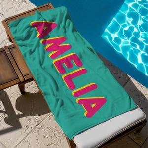 May include: A rectangular teal beach towel featuring the name AMELIA printed in large, bold, pink capital letters with a yellow drop shadow effect. The towel is laid out flat, displaying the vibrant color contrast and the centered text design.