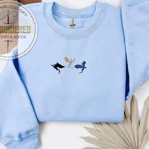 May include: A pale blue sweatshirt featuring three embroidered dragon designs in black, gold, and blue. The text "EMBROIDERED SWEATSHIRT & HOODIE" is visible on a circular label. The garment is folded, with a neutral-toned leaf arrangement in the corner.