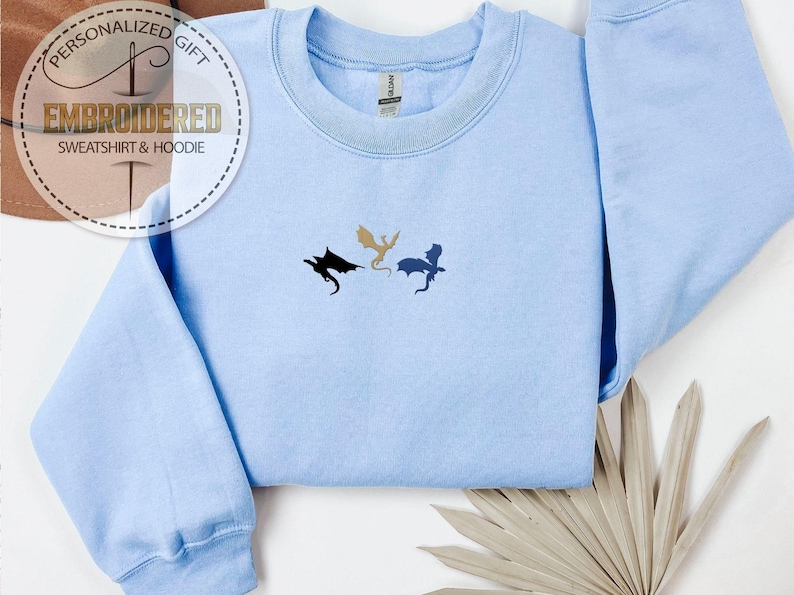 May include: A pale blue sweatshirt featuring three embroidered dragon designs in black, gold, and blue. The text "EMBROIDERED SWEATSHIRT & HOODIE" is visible on a circular label. The garment is folded, with a neutral-toned leaf arrangement in the corner.