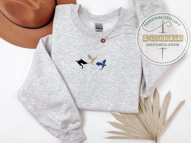 May include: A light gray sweatshirt with a crew neck and long sleeves. The sweatshirt features an embroidered design of three dragons in black, gold, and blue. A gold necklace with a circular pendant is on the sweatshirt. A round sticker on the right says 'Personalized Gift EMBROIDERED Sweatshirt & Hoodie'.