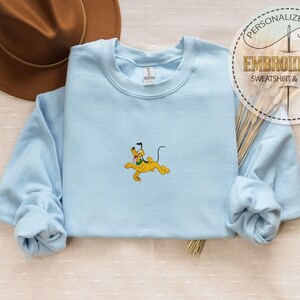Peut inclure: Un sweat-shirt bleu clair avec une broderie du personnage de dessin anim&eacute; Pluto en jaune. Le sweat-shirt a des manches longues et un col rond. Un chapeau marron est en arri&egrave;re-plan, et un autocollant circulaire indique "CADEAU PERSONNALIS&Eacute; SWEAT-SHIRT & HOODIE BROD&Eacute;."