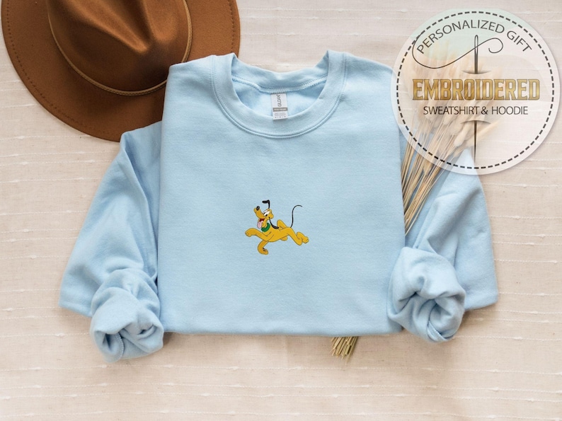 Peut inclure: Un sweat-shirt bleu clair avec une broderie du personnage de dessin anim&eacute; Pluto en jaune. Le sweat-shirt a des manches longues et un col rond. Un chapeau marron est en arri&egrave;re-plan, et un autocollant circulaire indique "CADEAU PERSONNALIS&Eacute; SWEAT-SHIRT & HOODIE BROD&Eacute;."