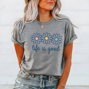 May include: Grey t-shirt featuring a daisy design with pink, yellow, and blue centres, and the text "life is good". The shirt is a casual item of clothing, suitable for everyday wear.