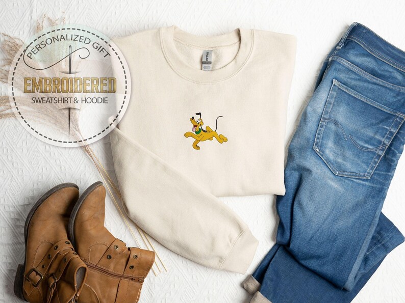 Peut inclure: Sweat-shirt cr&egrave;me brod&eacute; avec un dessin de Pluto. L'image comprend un jean bleu et des bottes marron. Le texte sur l'image dit "CADEAU PERSONNALIS&Eacute; SWEAT-SHIRT & HOODIE BROD&Eacute;".